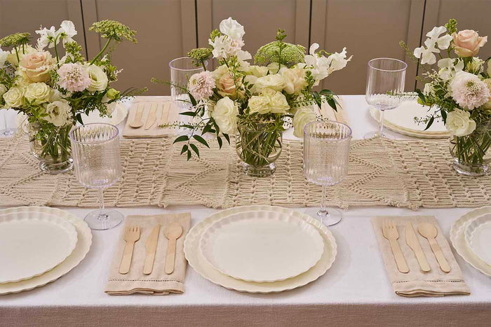 A table set for four with Sophistiplate Simply Eco Dinner Plate in white, beige napkins, wooden cutlery, textured glasses, and centerpieces of white and pale pink flowers in glass vases on a woven table runner.
