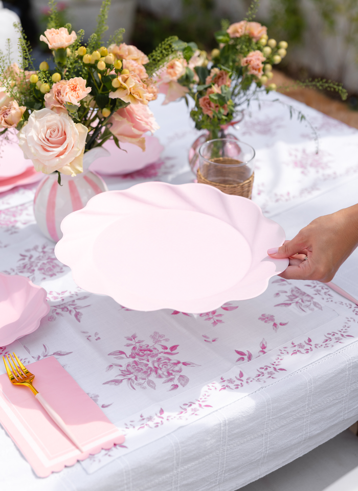 A hand holds a large blush compostable plate from the Eco-Friendly Table Setting-Blush by Sophistiplate LLC over a table with matching pink plates, gold cutlery, floral napkins, and vases of blush roses on a white floral tablecloth.