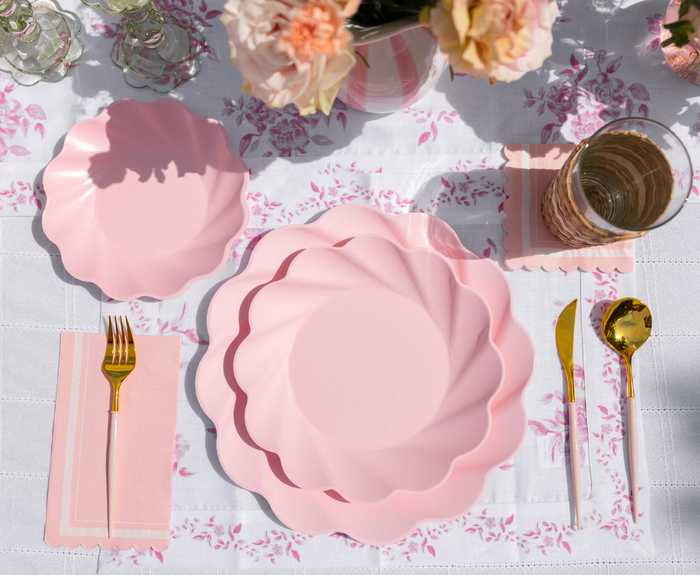 The Eco-Friendly Table Setting- Blush by Sophistiplate LLC features pink scallop plates, gold cutlery on a pink napkin, a glass with a pink coaster, and a floral tablecloth with a vase of pink flowers for a soft, elegant look.