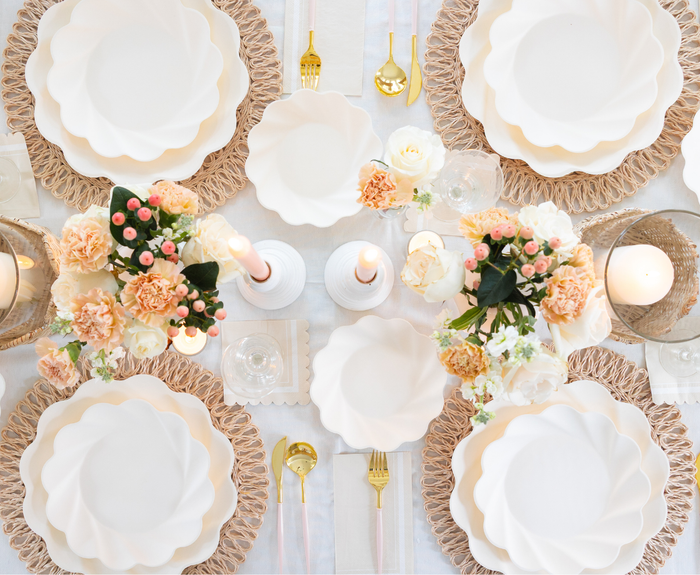 An elegant table setting with Sophistiplate Simply Eco Extra Large Plates in cream, gold cutlery, woven placemats, peach and white floral centerpieces, and candles on a light tablecloth.