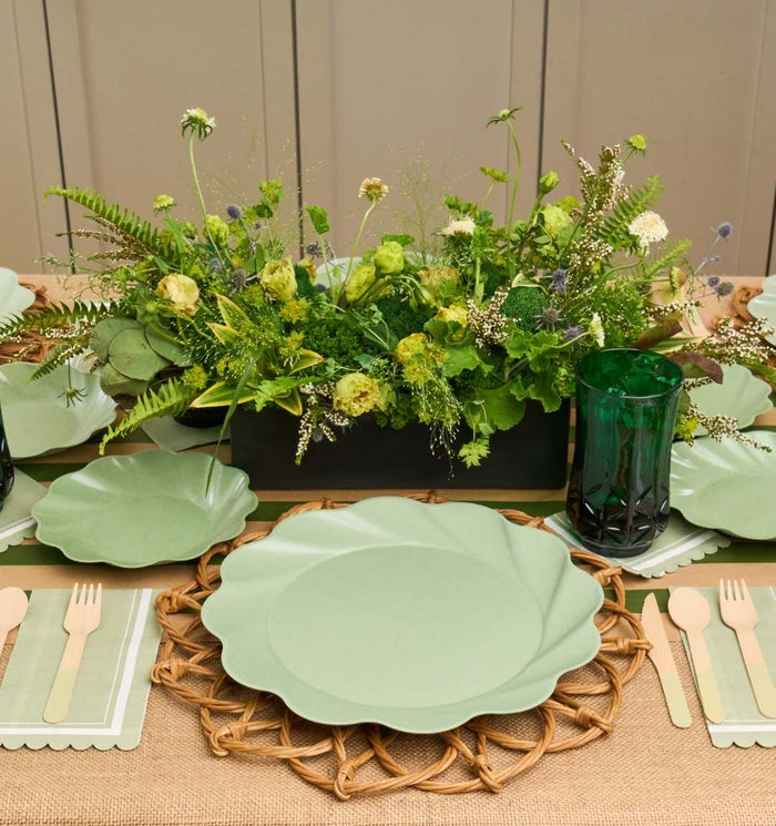 A beautiful table set with Sophistiplate LLC’s Eco-friendly Table Setting- Sage, featuring light green scalloped plates, wooden cutlery, green glasses, and a centerpiece of assorted foliage on a burlap cloth and woven placemats.