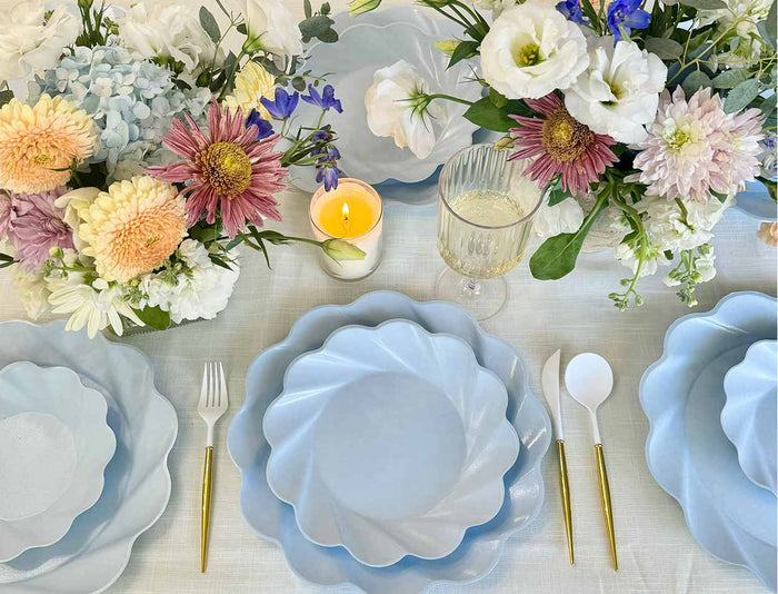A beautifully set table with Sophistiplate Simply Eco Extra Large Sky Blue Plates, gold and white cutlery, pink, white, and purple floral arrangements, a lit candle, a glass of white beverage, and a light tablecloth.