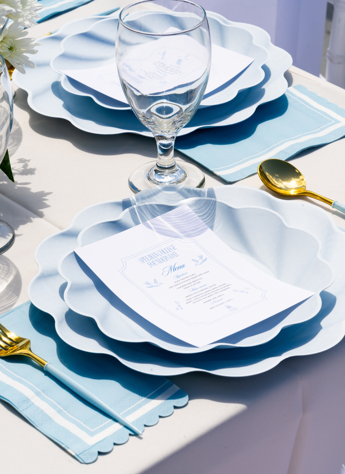 The Sophistiplate Eco-Friendly Table Setting - Sky Blue features layered plates, gold cutlery, blue napkins, a wine glass, and a printed menu on each plate atop a white tablecloth in bright natural light.
