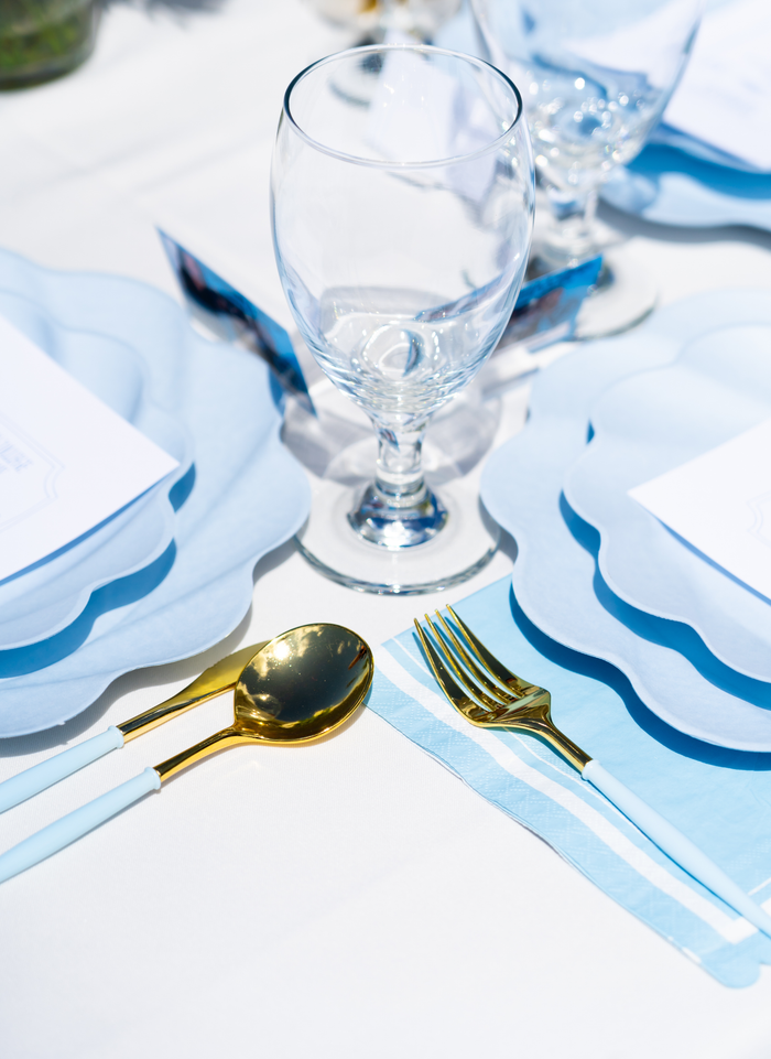 A close-up of the Sophistiplate Eco-Friendly Table Setting - Sky Blue, showcasing blue scalloped plates, a clear wine glass, and gold cutlery with blue handles on a white tablecloth.