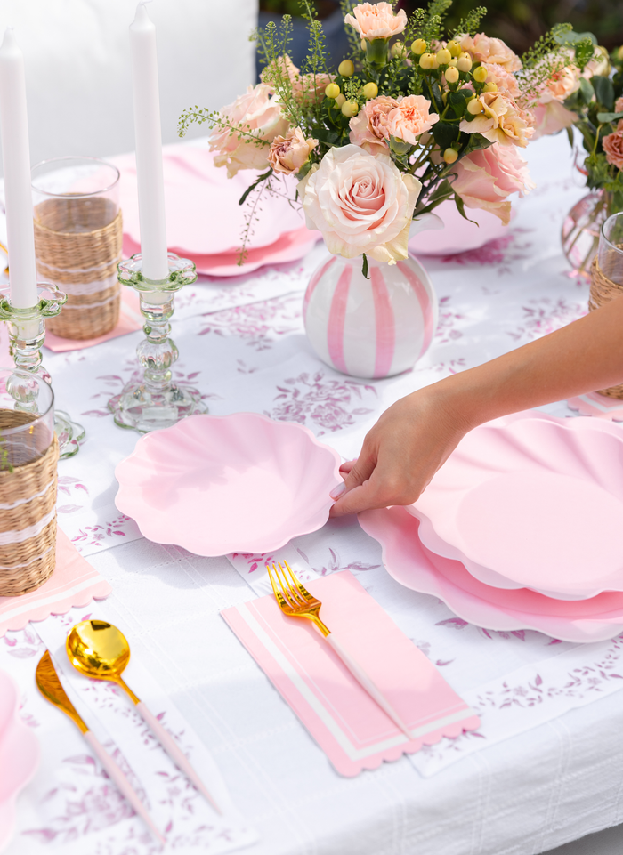 A hand sets a Sophistiplate LLC Eco-Friendly Table Setting- Blush plate with scalloped pink edges on a floral tablecloth, surrounded by gold cutlery, pink napkins, candlesticks, and vases of blush roses and greenery.