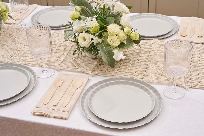 A table set with Sophistiplate’s Simply Eco Dinner Plate-Concrete, wooden cutlery on a napkin, clear ribbed glasses, a woven beige runner, and a white-green floral centerpiece completes the eco-friendly tablescape.