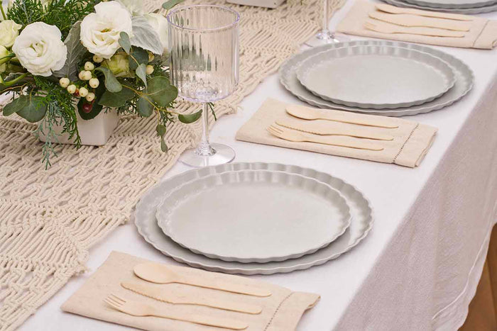 A close-up of a dining table with Sophistiplate’s Simply Eco Dinner Plate-Concrete, beige napkins with wooden cutlery, clear ribbed glasses, and white flowers and greenery arranged on a woven table runner.