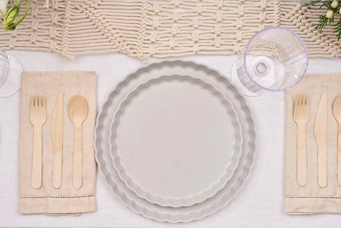 Overhead view of a neutral table setting with two stacked Sophistiplate Simply Eco Dinner Plates in Concrete (8/pkg), beige cloth napkins, wooden cutlery, clear glasses, and a textured runner atop a white tablecloth.