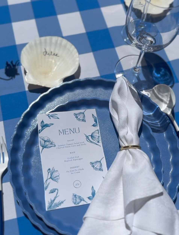 A blue and white checkered tablecloth set with a Sophistiplate Simply Eco Salad Plate in French Blue, a white napkin, floral menu, wine glass, silverware, and a shell place card handwritten with "Julie.