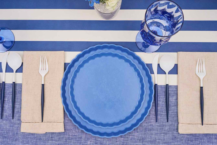 A top-down view of Sophistiplate's Eco-Friendly Table Setting- French Blue features two stacked compostable plates, beige napkins, white plastic flatware with dark handles, and a blue glass on a blue and white striped tablecloth.