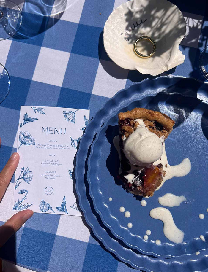 A slice of pie with ice cream is served on a Sophistiplate Simply Eco Salad Plate- French Blue (8/pkg), set on a blue-and-white checkered table beside a floral menu, gold ring in a shell dish, and a hand in sunlight.
