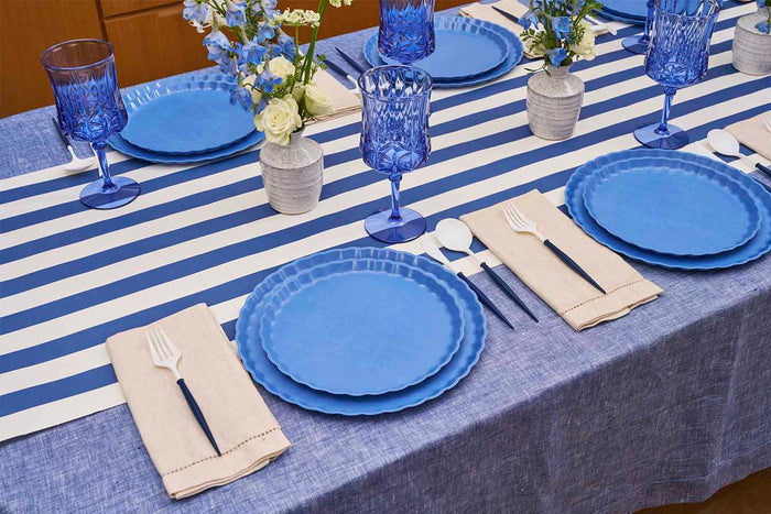 The Eco-Friendly Table Setting- French Blue by Sophistiplate features blue glasses, beige napkins with white cutlery, blue and white floral vases, and a blue tablecloth with a blue and white striped runner.