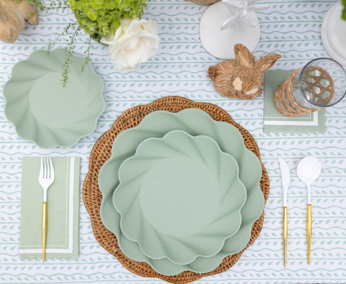 A Sophistiplate LLC Eco-friendly Table Setting- Sage features pastel green scalloped plates on a woven placemat, gold and white cutlery on a napkin, a glass, and a decorative bunny on a floral-patterned tablecloth.