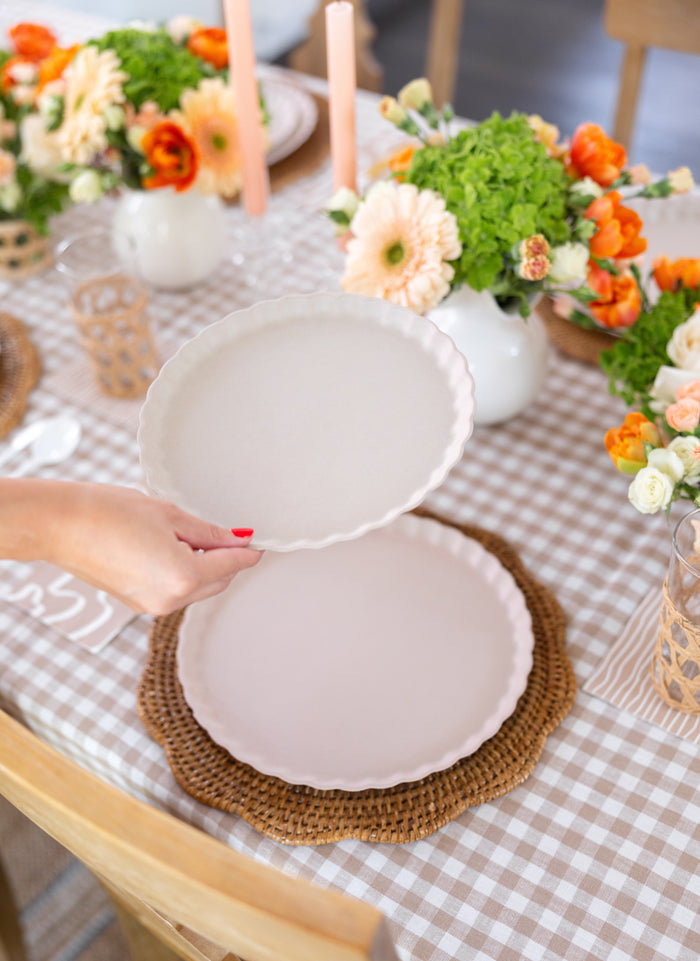 A person with red-painted nails sets a Sophistiplate Eco-Friendly Table Setting- Sand plate on another atop a woven placemat, surrounded by a checkered tablecloth, floral arrangements, candles, and sustainable tableware.