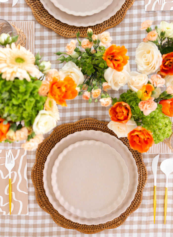 A top-down view of a table set with white plates on woven placemats, orange and white flowers, a beige checkered cloth, gold utensils, and Sophistiplate Guest Towel Eco Sand paper napkins for a stylish touch.