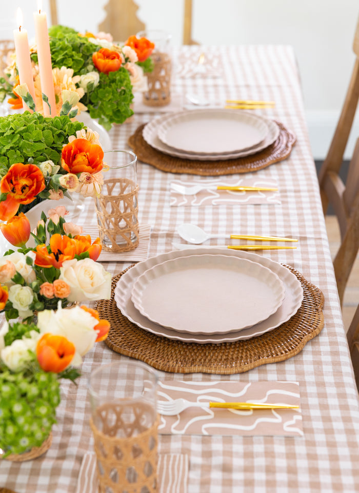 A spring table features a beige gingham cloth, woven placemats, cream plates, gold-white cutlery, wicker-wrapped glasses, Sophistiplate Guest Towel Eco Sand napkins, and vases with orange, white & green flowers.