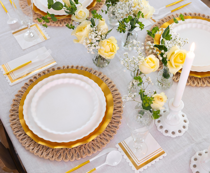 The Eco White & Gold Table Setting by Sophistiplate LLC features eco-friendly party plates, gold cutlery, clear glasses, gold-striped white napkins, a white candle, yellow roses, and small white flowers on a crisp white tablecloth.