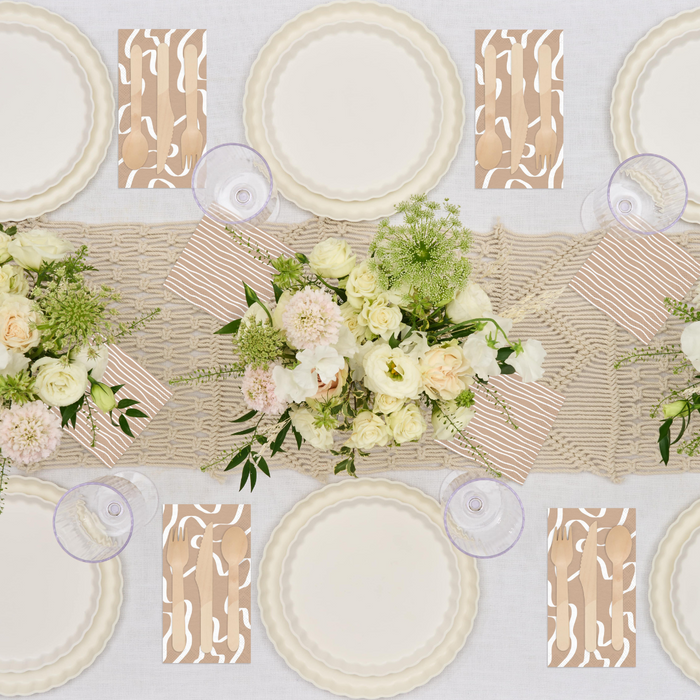 A tablescape featuring Sophistiplate Simply Eco Salad Plates in white, clear glasses, patterned napkins with wooden cutlery, and a central garland of white and pale peach flowers with greenery on a woven runner.