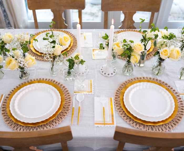 The Eco White & Gold Table Setting by Sophistiplate LLC sets a table for four with designer disposable plates, yellow and woven placemats, gold cutlery, white napkins, yellow roses with greenery in vases, and two lit white candles as the centerpiece.