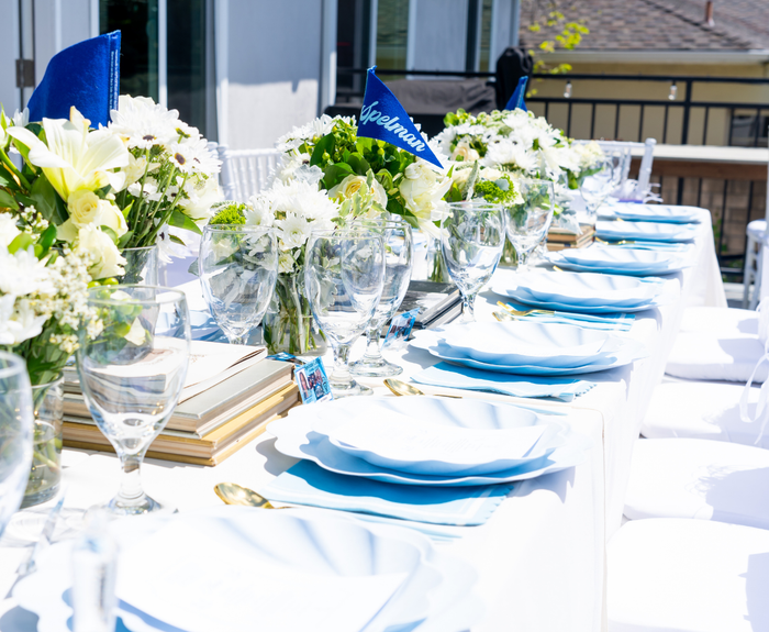 An elegantly set outdoor table features Sophistiplate's Simply Eco Compostable Table Setting in Sky Blue, glassware, white floral centerpieces, and blue graduation caps as decor, all glowing in the sunlight.