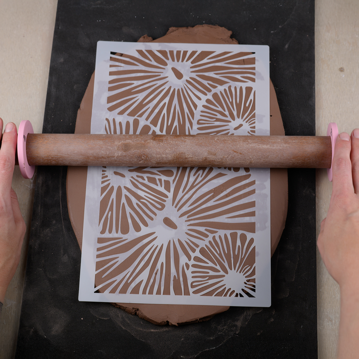 Person using a rolling pin with a decorative stencil to imprint patterns on clay.