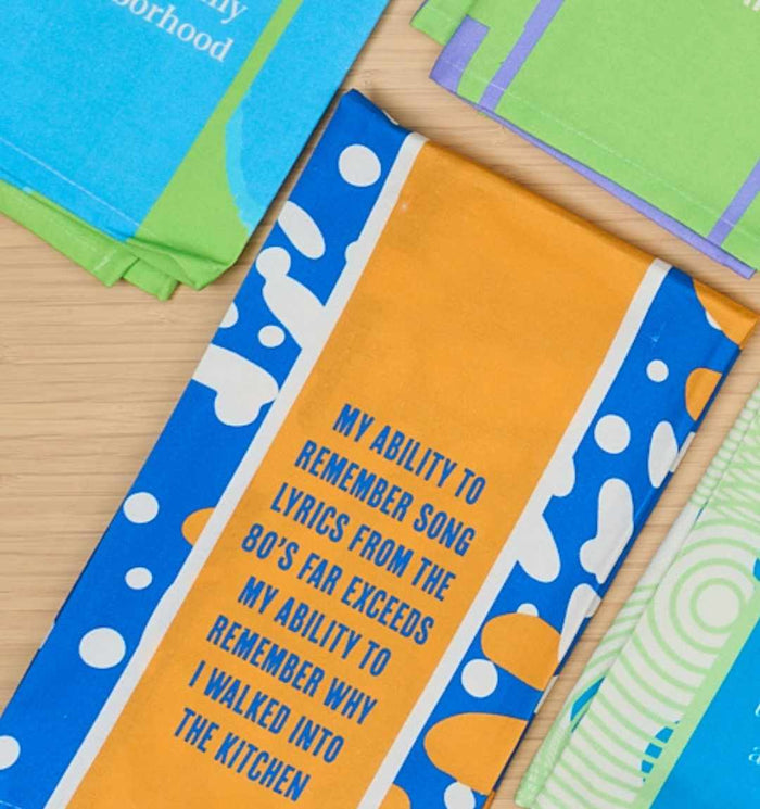 The Drinks On Me Song Lyrics Dish Towel features blue, orange, and white designs with the phrase: My ability to remember 80s song lyrics far exceeds my ability to remember why I walked into the kitchen. Other towels are partly visible.