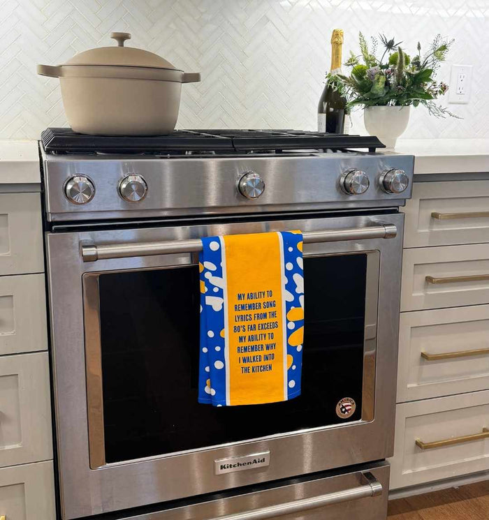 A modern stainless steel oven with a beige pot on top and a Drinks On Me Song Lyrics Dish Towel on the door. The towel reads, “My ability to remember song lyrics from the 80’s far exceeds my ability to remember why I walked into the kitchen.” Flowers and champagne nearby.
