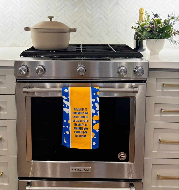 A modern stainless steel oven displays the Song Lyrics Dish Towel by Drinks On Me, featuring witty text and bold yellow-blue patterns. A beige pot sits on the stove, with flowers nearby—making it a fun and quirky housewarming gift.