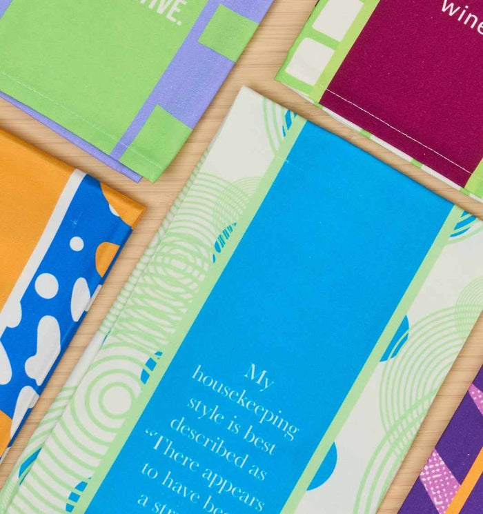 Colorful patterned fabric pieces are displayed on a light wood surface. The Drinks On Me Struggle Dish Towel features modern geometric designs and the funny phrase “My housekeeping style is best described as: There appears…”—perfect for kitchen humor.