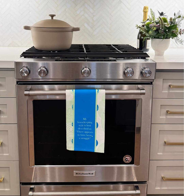 A stainless steel oven with a beige pot on the stove and a Drinks On Me Struggle Dish Towel in blue and green hanging from the door. Champagne and flowers on the white countertop make this scene perfect for a housewarming gift.