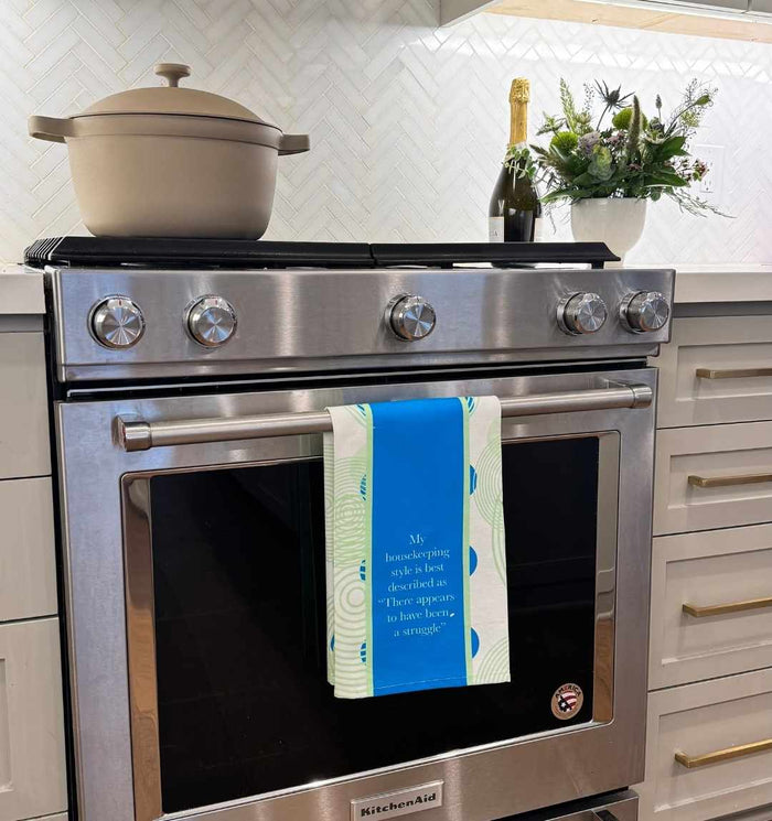 A modern stainless steel oven with a cream pot, champagne bottle, and flowers on top. The Drinks On Me "Struggle Dish Towel" hangs from the handle—a blue and white towel that adds kitchen humor, perfect for housewarming. White cabinets, herringbone backsplash.