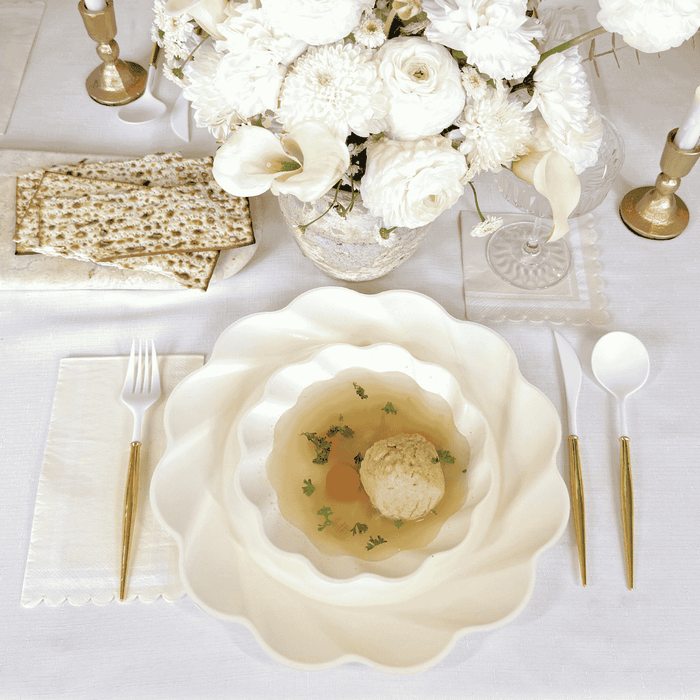 The Stunning Seder Table Setting by Sophistiplate includes designer disposable plates, gold cutlery, matzo ball soup, stacked decorative plates, matzo crackers, and a vase of white flowers on a crisp white tablecloth.