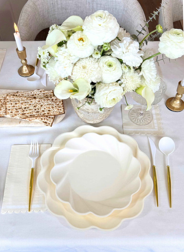 The Stunning Seder Table Setting by Sophistiplate features scalloped white disposable plates, gold and white cutlery, folded napkins, matzo crackers, a white floral centerpiece, candles, and a crisp white tablecloth.