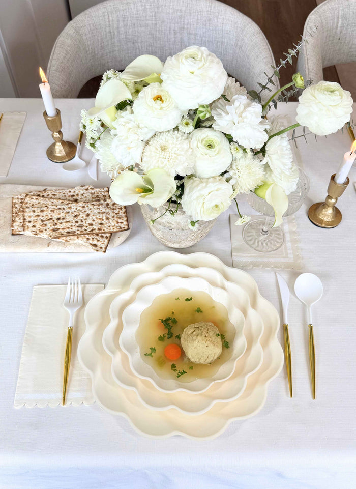 The Stunning Seder Table Setting by Sophistiplate features gold cutlery, designer disposable plates, matzo ball soup with carrots, matzo crackers, a white floral centerpiece, and two lit candles in gold holders for an elegant display.