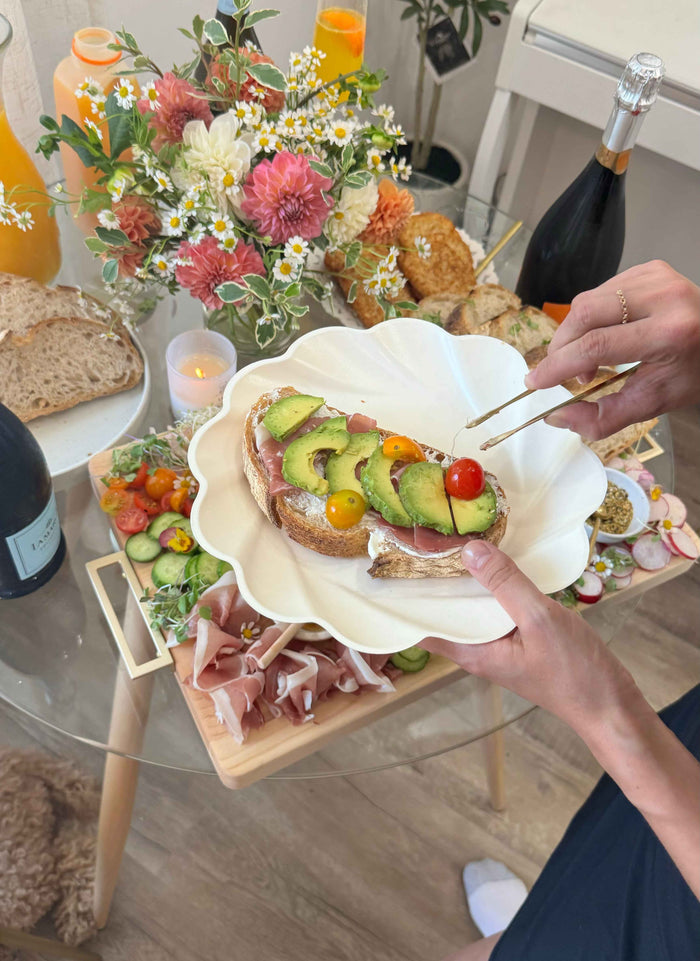 A person garnishes avocado toast on a Sophistiplate Stunning Seder Table Setting, adding style to a tablescape-in-a-box with flowers, wine, juice, bread, prosciutto, and more brunch treats.