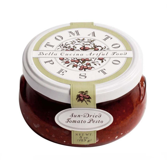 A 6 oz glass jar of Bella Cucina Sun-dried Tomato Pesto with a white lid and floral label, made with extra virgin olive oil for rich flavor in every spoonful.