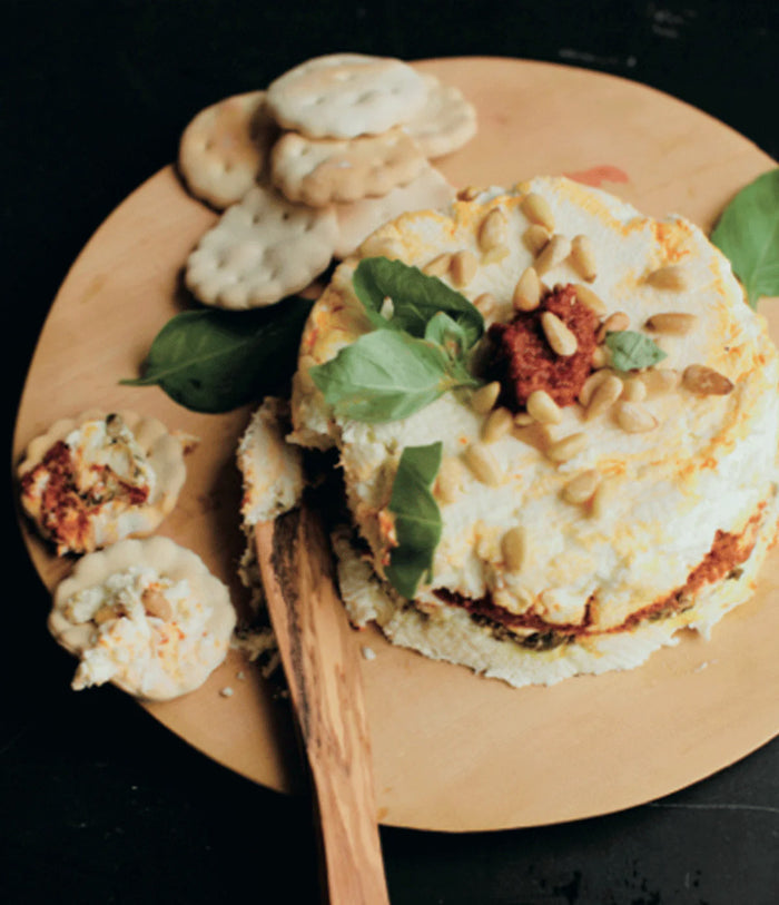 Bella Cucina Sun-dried Tomato Pesto spread is served in a round, layered style with Parmesan, garnished with basil and pine nuts on a wooden board surrounded by crackers. A wooden knife with cheese rests nearby.