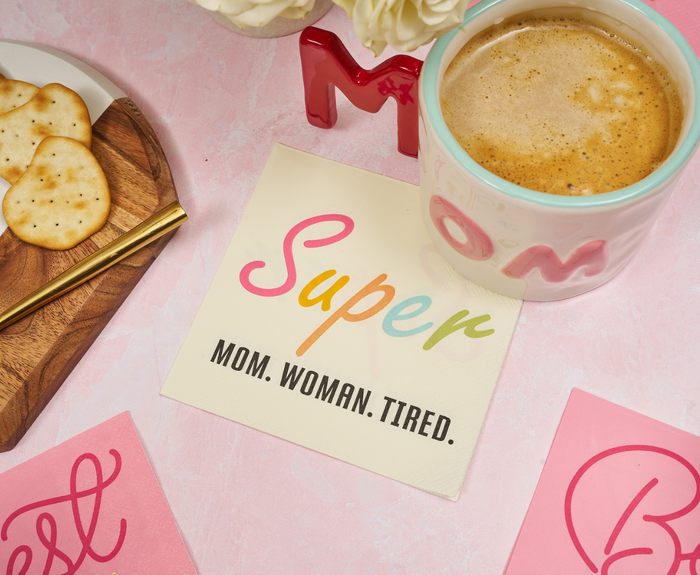 Coffee and crackers rest on a wooden tray alongside Sugarplum Super Mom Woman Tired Cocktail Napkins (26pk), set on a pink floral surface—perfect Mother’s Day party supplies.