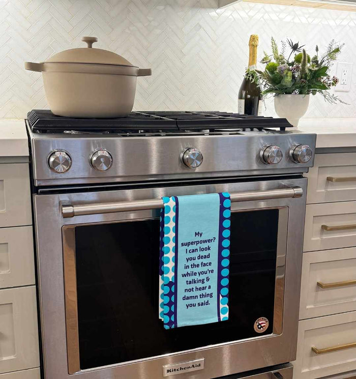 A beige pot sits atop a modern stove, while the Drinks On Me Superpower Dish Towel with polka dots hangs on the oven door, featuring the quote: "My superpower? I can look you dead in the face while you’re talking & not hear a thing.