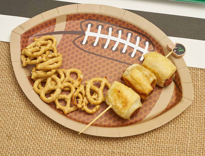 The Sophistiplate Tailgate Die Cut Football Salad Plates (8pk) display a handful of mini pretzels and three pigs in a blanket on a skewer, set atop a textured brown table covering.