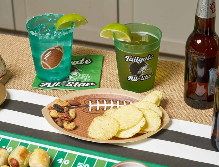 Two drinks with lime wedges, football-themed napkins, and Sophistiplate Tailgate Die Cut Football Salad Plates filled with chips and nuts are displayed on a festive table. Game day tableware and a beer bottle create the ideal sports party setup.