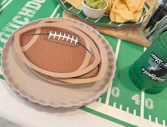 Sophistiplate Tailgate Die Cut Football Salad Plates (8pk) stacked on a football field tablecloth with chips, guacamole, and a green cup create the perfect game day tableware setup for your next celebration.