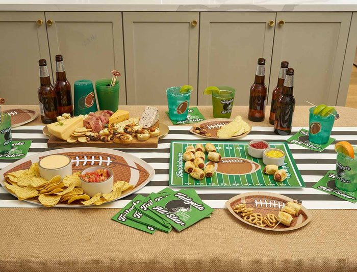A table set for a football party with Sophistiplate Tailgate Die Cut Football Salad Plates (8pk), game day tableware, chips, cheese, sliders, drinks in green cups and bottles, plus themed napkins and trays.