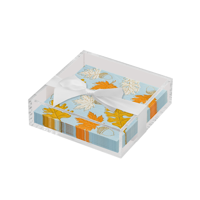 The Sophistiplate Thanksgiving Cocktail Napkin with Napkin Tray holds festive napkins featuring a blue background with orange, yellow, and white leaf designs, tied with a white ribbon—ideal for a modern tabletop.