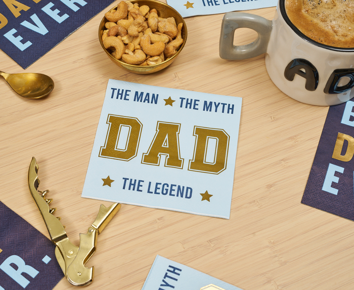 A set of Sugarplum's The Man The Myth Cocktail Napkins (26pk) is shown on a wooden surface with a DAD mug, nuts in a bowl, golden spoon, cards, Best Dad Ever napkins, and a golden corkscrew.