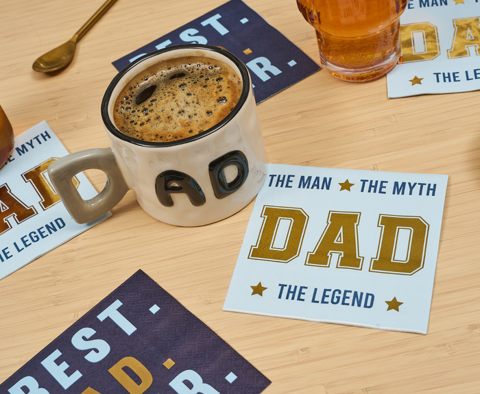 A coffee mug labeled DAD sits on a wooden table next to Sugarplum’s The Man The Myth Cocktail Napkins (26pk), which read THE MAN THE MYTH DAD THE LEGEND in bold letters, with other napkins and a drink nearby.