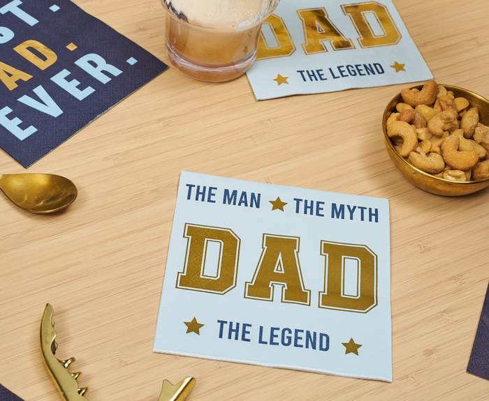 Sugarplum’s The Man The Myth Cocktail Napkins (26pk) feature “THE MAN THE MYTH DAD THE LEGEND” in bold letters, perfect for Father’s Day celebrations. Shown on a wooden table with snacks and drinks nearby.