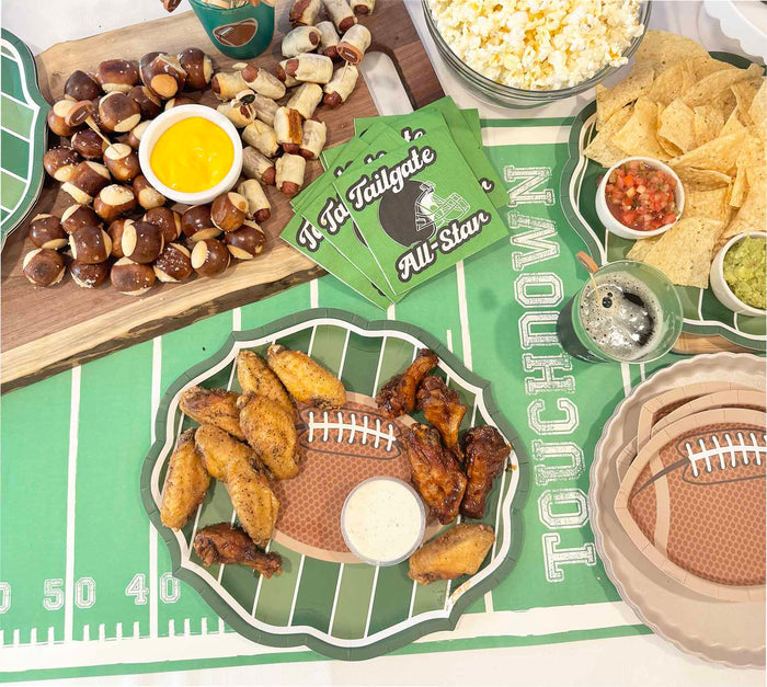 The Superbowl Set by Sophistiplate showcases chicken wings, pretzel bites with cheese dip, popcorn, tortilla chips with salsa and guacamole, drinks, designer disposable plates, and football-shaped napkins on a green field tablecloth.