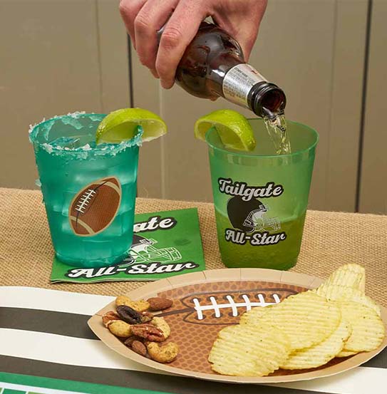A hand pours a drink into a green cup from The Superbowl Set by Sophistiplate, featuring "Tailgate All-Star" and football designs. Designer disposable plates hold chips and nuts on a football field-themed table—perfect for parties.