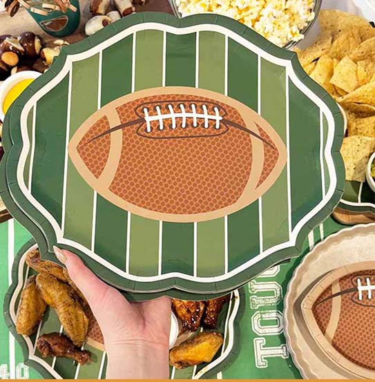A hand holds "The Superbowl Set" by Sophistiplate—designer football-shaped plates with a field design—surrounded by snacks, ideal for parties or any football-themed gathering.